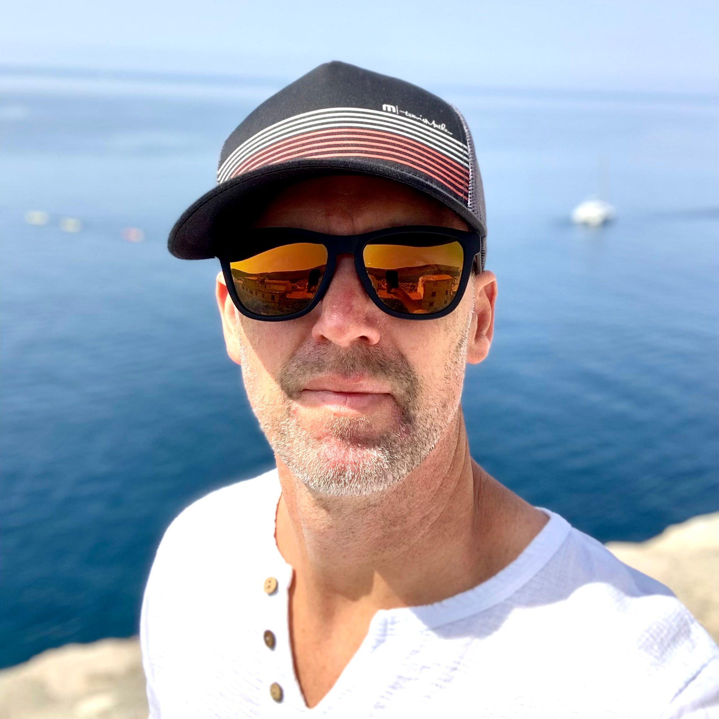 Man looking at camera wearing cap and sunglasses with ocean behind him.