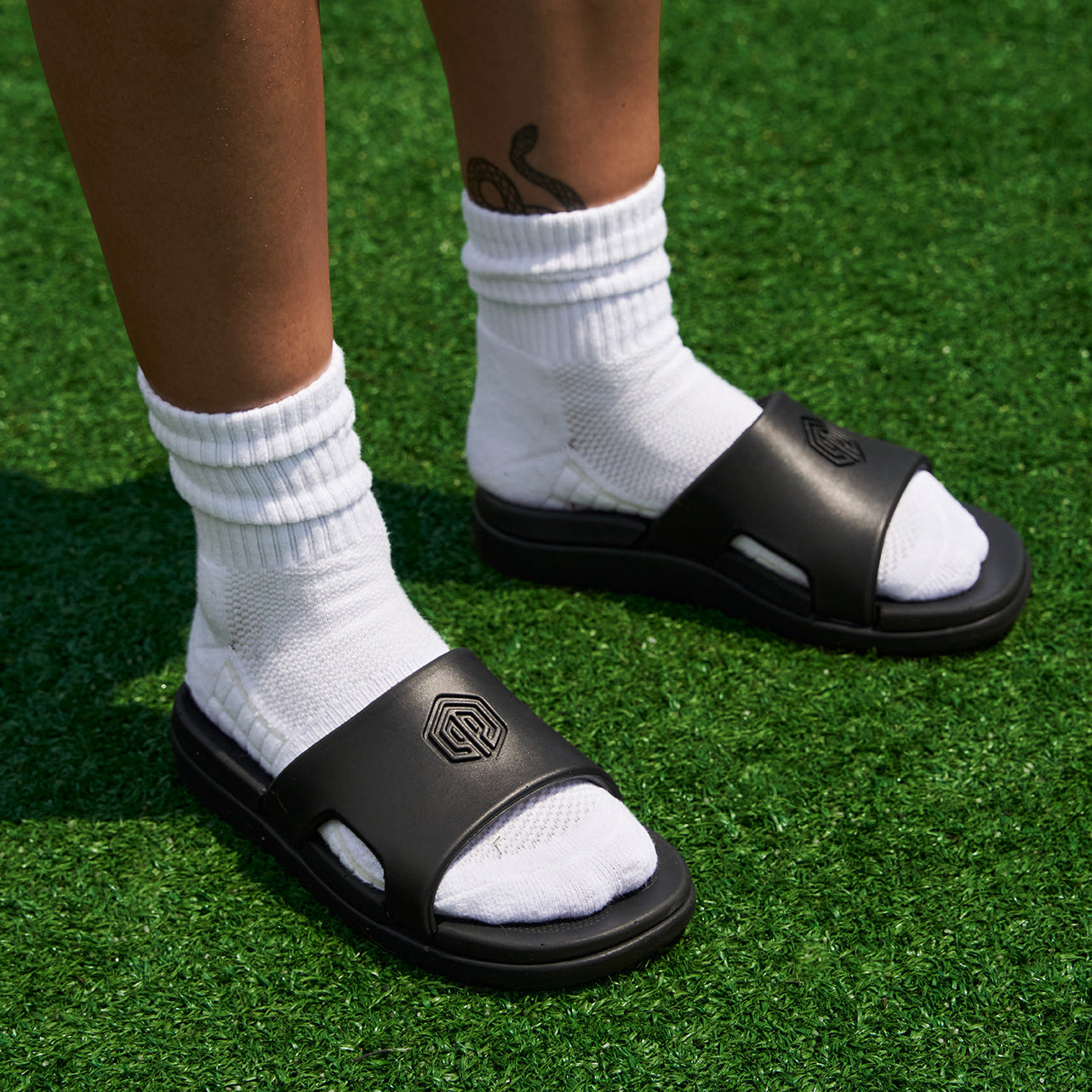 Two feet wearing white socks and black slides on green artificial grass.