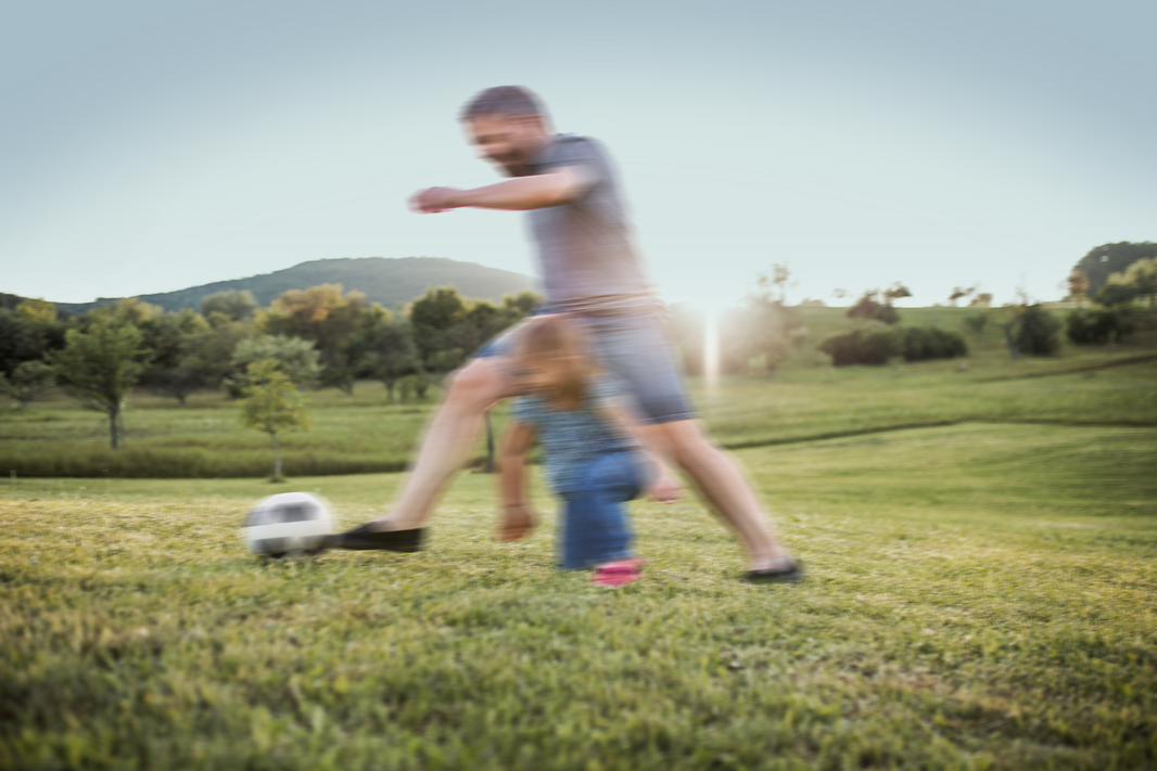 Man playing with young girl in grassy field kicking a soccer ball.