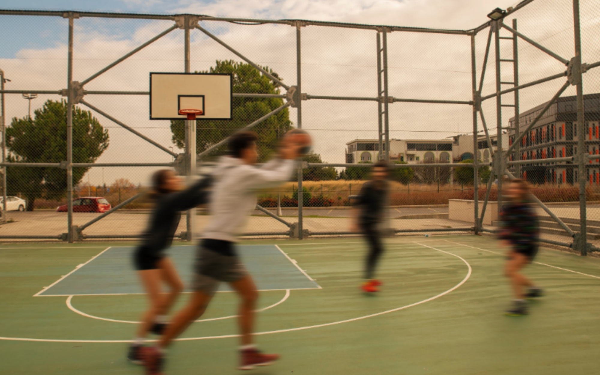 Blurred image of people playing basketball on an outdoor court with a building in the background.