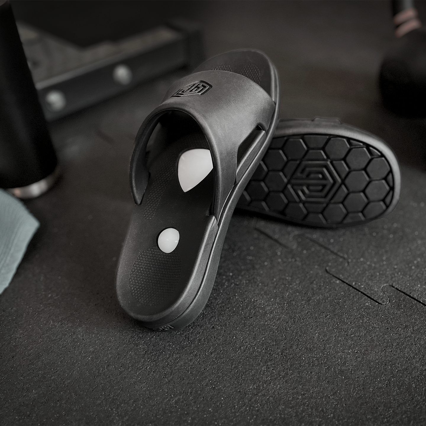 Black slide sandals on a dark textured surface with gym equipment in the background