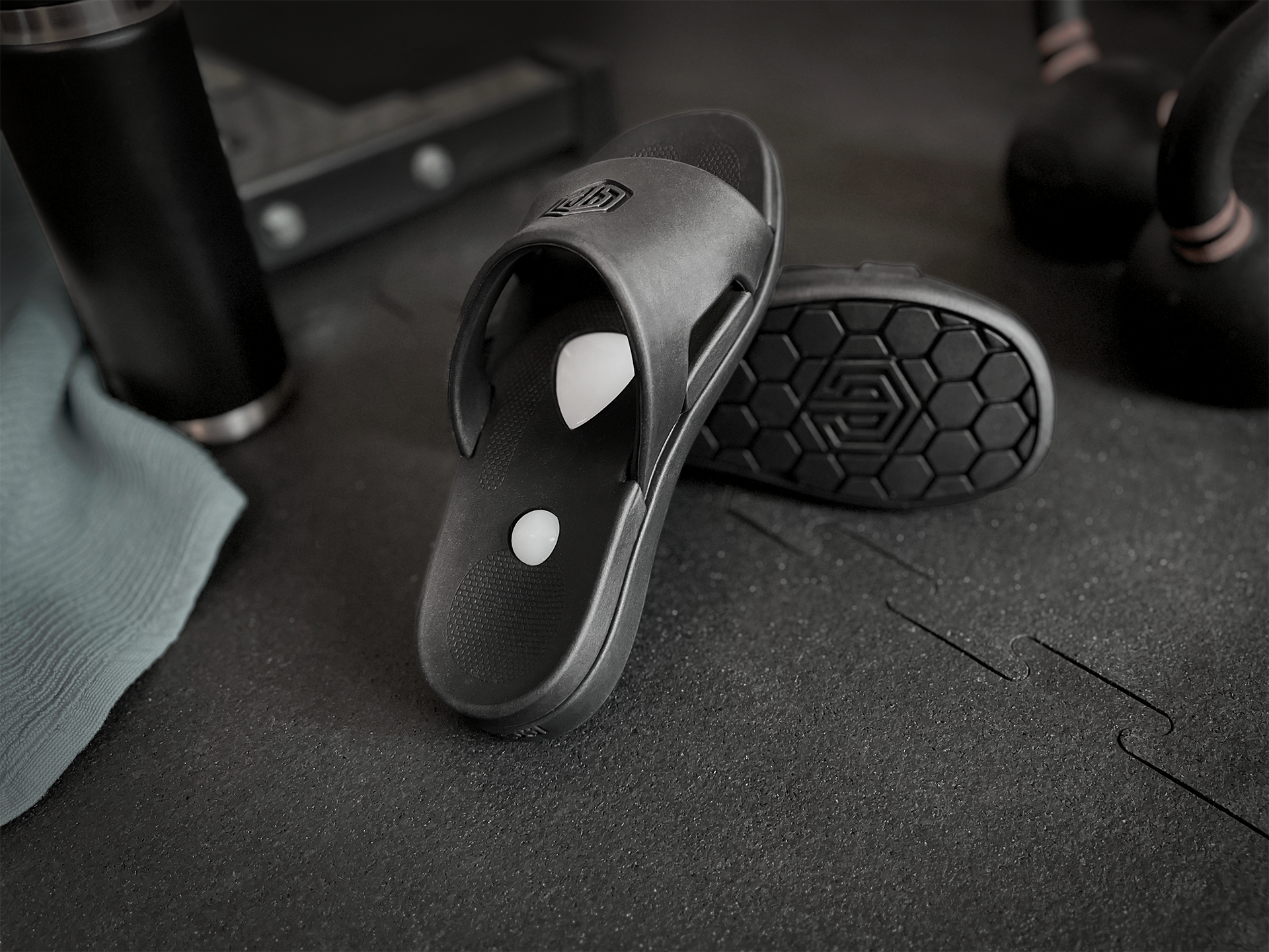 Black slide sandals on a dark textured surface with gym equipment in the background
